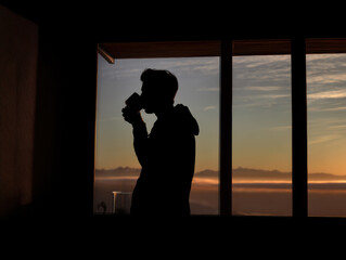 Silhouette person drinking coffee by large window at sunrise with warm sky and mountain view evoking calm morning reflection