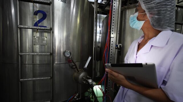 Industrial Quality Control: A female technician wearing a lab coat and mask inspects large stainless steel tanks in an industrial setting, holding a tablet for data recording.