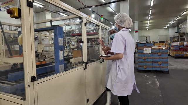 Food Production Line: Two workers in sterile lab coats and hairnets monitor a high-tech food processing machine on a factory floor, ensuring quality and efficiency.