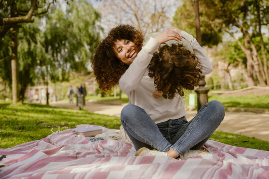 A mother sits on a blanket and laughs while holding her son on her shoulders