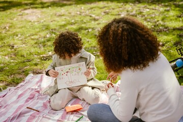 The son shows the drawing to his mother, who is sitting next to him on the blanket