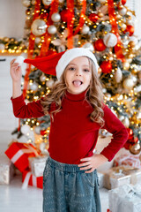 a child girl in a Santa Claus hat rejoices in the new year and presents at the Christmas tree with a red decor at home, a happy child receives gifts