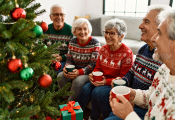 Senior friends in festive sweaters enjoying hot drinks and conversation by Christmas tree with decorations and presents in cozy living room during holiday season
