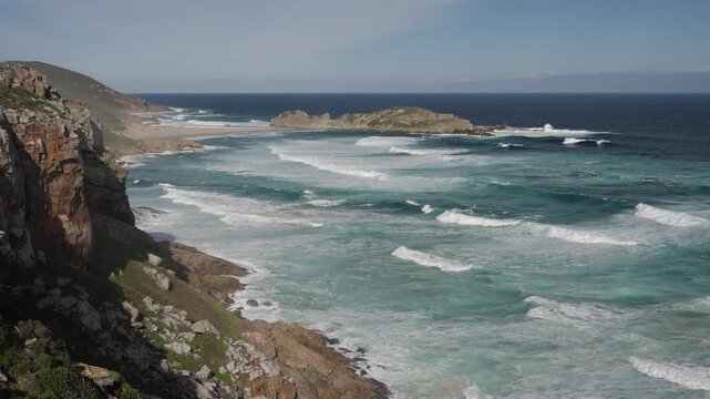 Breathtaking 4K 30fps video of Robberg Nature Reserve, Plettenberg Bay, Garden Route, South Africa. Rugged cliffs meet the Indian Ocean under a vivid blue sky with streaking clouds, showcasing nature.