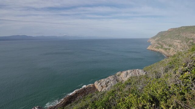 Breathtaking 4K 30fps video of Robberg Nature Reserve, Plettenberg Bay, Garden Route, South Africa. Rugged cliffs meet the Indian Ocean under a vivid blue sky with streaking clouds, showcasing nature.