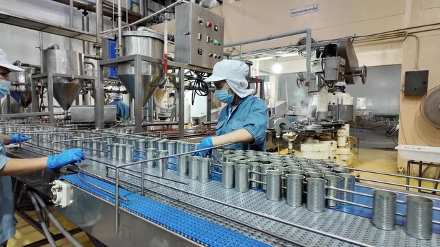 Several metal cans are lined up on a conveyor in a production facility. Workers in the background monitor the machinery's operation, ensuring a smooth flow of the manufacturing process.