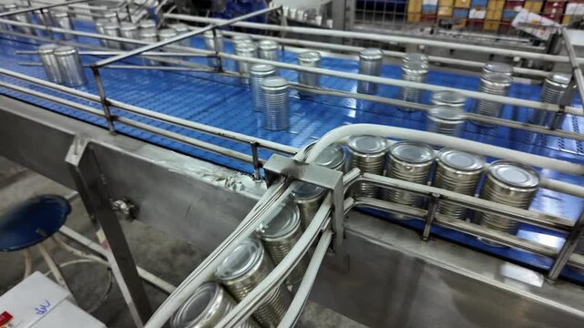 Several metal cans are lined up on a conveyor in a production facility. Workers in the background monitor the machinery's operation, ensuring a smooth flow of the manufacturing process.