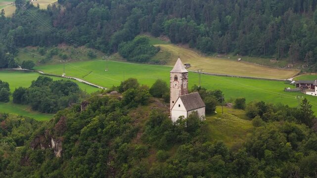 Chiesa di Santa Verena positioned on a tree-covered rise, aerial view. Dolomites countryside with alpine terrain