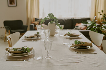 Dining table set with glassware, plates, cutlery, and napkins decorated with green leaves, positioned in living room with sofa and plants visible in background