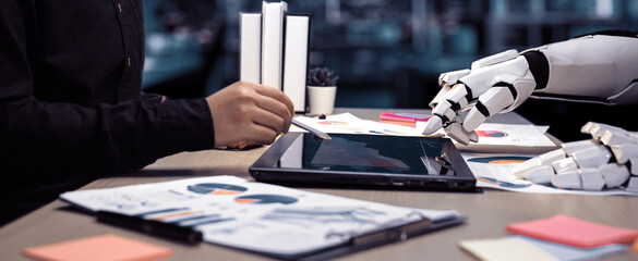 A futuristic office scene showcasing a humanoid robot hand engaged with a tablet, collaborating with a human colleague amidst charts and notes, symbolizing innovation.Noogenesis
