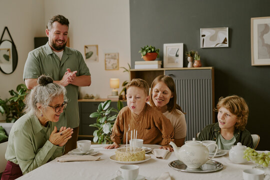 Caucasian child blowing out candles on birthday cake surrounded by family including mother, grandmom, father and brother clapping