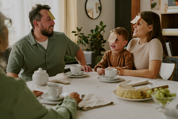 Caucasian man, Caucasian woman, and Caucasian child sitting at dining table playing guessing game with sticky notes on foreheads, smiling and laughing together during family breakfast