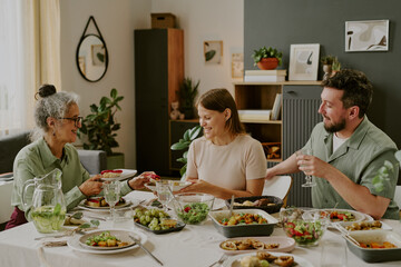 Senior Caucasian woman, young adult Caucasian woman, and young adult Caucasian man sitting at dining table sharing meal, smiling and interacting with each other, surrounded by food