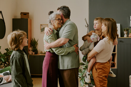 Middle aged Caucasian man hugging senior mother while his wife holding child with stuffed animal and another child standing nearby, family gathering indoors - Powered by Adobe