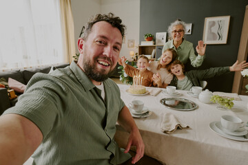 Portrait of Caucasian young adult man taking selfie with smiling Caucasian senior woman, Caucasian young adult woman, two Caucasian children celebrating birthday at dining table