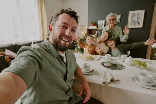 Portrait of Caucasian young adult man taking selfie with smiling Caucasian senior woman, Caucasian young adult woman, two Caucasian children celebrating birthday at dining table