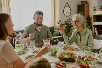Caucasian middle aged man, Caucasian young adult woman, and senior woman sitting at table eating healthy meal together, smiling and interacting during family gathering