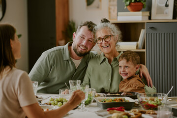 Caucasian middle aged man, senior Caucasian woman, and Caucasian child sitting together at dining table, smiling and hugging while enjoying meal with another woman visible from behind