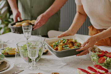 Caucasian young woman serving vegetable casserole while Caucasian young man holding basket of bread, both arranging food on dining table with fresh salad and glassware visible