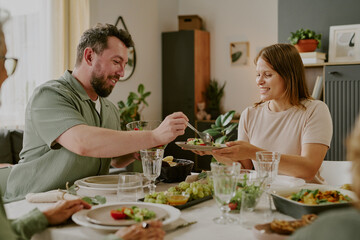 Caucasian middle aged man serving food to Caucasian young adult woman at dining table, both smiling and interacting during shared meal, other people partially visible around table