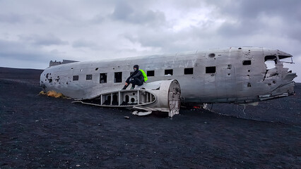 A lone male traveler wearing a face mask and bright jacket sits on the wing of the wrecked DC-3, a modern explorer contemplating the desolate beauty of the Sólheimasandur black sand beach in Iceland.