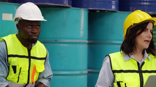 Warehouse Guardians: Two workers, a man and a woman wearing hard hats and safety vests, stand in a warehouse amidst large blue barrels, overseeing operations and ensuring safety.
