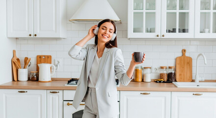 A beautiful young woman, smiling, dancing while enjoying music with her headphones and holding a coffee mug in her kitchen