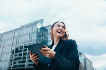 Confident young woman smiling while holding a tablet outdoors in a modern urban environment showcasing technology and communication mock up