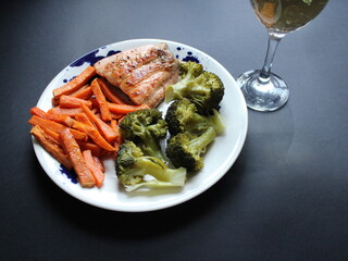 Grilled salmon, steamed potatoes and broccoli on a white plate and a glass of white wine on dark background