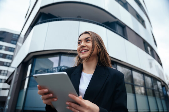 Young professional woman smiling while holding tablet outdoors in urban setting with modern architecture showcasing technology and lifestyle concept