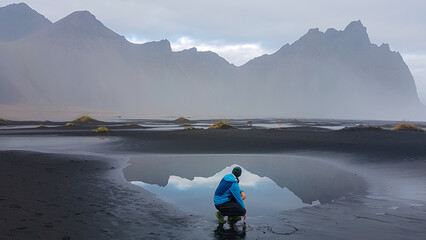 A man crouches by a tidal pool on Stokksnes black sand beach, drawing a heart in the sand with the...