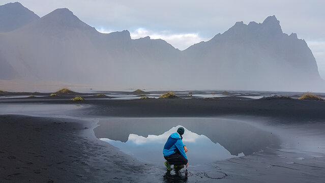 A man crouches by a tidal pool on Stokksnes black sand beach, drawing a heart in the sand with the majestic, misty Vestrahorn mountain perfectly reflected in the tranquil water beside him in Iceland.