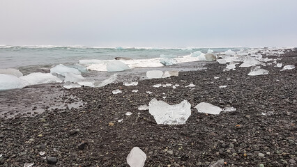 Chunks of clear glacial ice, like diamonds, are scattered across pebbly black sand of Iceland's famous Diamond Beach, with ocean waves washing ashore under moody, overcast sky, unique natural wonder