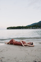 Relaxed woman lying on the sandy beach near calm sea water with distant green hills and soft pastel sky at sunset.