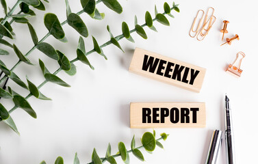 Wooden blocks with the words weekly report on white background with green leaves, pens and office supplies