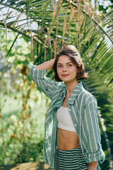 Portrait of a young woman outdoors in a striped green shirt and white crop top amid lush tropical...