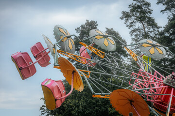 Bright pink and yellow carnival ride swings high in the air, with flower-shaped tops spinning against a blue sky and trees in the background.