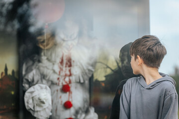 A young boy in a gray hoodie stares at a menacing clown figure with bloodstained costume and red balloons inside a glass display, creating a chilling horror-themed scene.