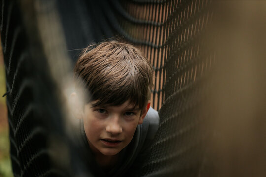 Smiling boy in a gray hoodie gives a thumbs-up while lying inside a black rope net tunnel at an outdoor play area.