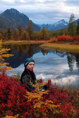 A young female hiker enjoys the views of the autumn taiga. A traveler is relaxing near a lake that displays snow-capped mountains. A forest painted with the bright colors of the changing season