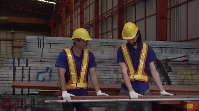 Warehouse Workers in Action: Two industrious workers, clad in hard hats and high-visibility vests, collaborate while handling long metal beams in a bustling warehouse setting.
