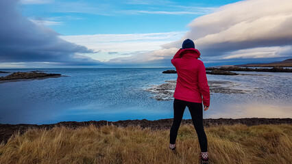 A female traveler in a pink jacket, seen from behind, stands on a grassy shore, gazing out at the serene, blue sea and dramatic sky on the beautiful Snaefellsnes peninsula, Iceland, lost in thought.