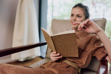 Young woman in casual brown outfit reading a book in a cozy indoor setting, evoking relaxation and...