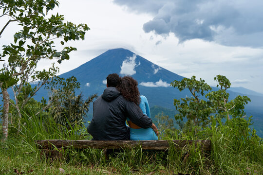 A couple of lovers are sitting on a bench, hugging each other and enjoying the view of the popular sacred cloud-covered Mount Agung on the island of Bali.
