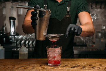 Bartender pouring red cocktail into glass with ice and strainer