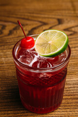 Refreshing red cocktail garnished with cherry and lime on wooden table