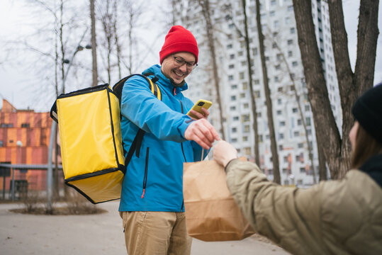 Smiling delivery man with yellow insulated backpack giving paper bag with takeaway food to customer and using smartphone app for contactless payment in city street