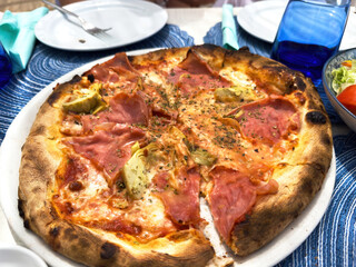 Wood fired pizza with ham, artichokes, cheese and herbs on a white dish outdoors, shared at a sunny table during a relaxed Mediterranean family lunch.