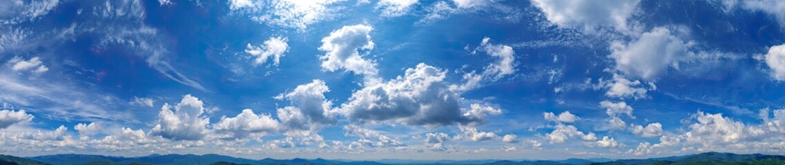 Panoramic view of a vibrant blue sky filled with fluffy white clouds. A hazy line of distant hills or mountains hints at a landscape below