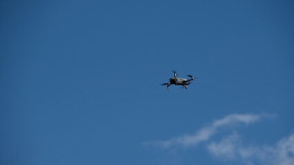 Small drone captured in flight against a clear blue sky with a few clouds.